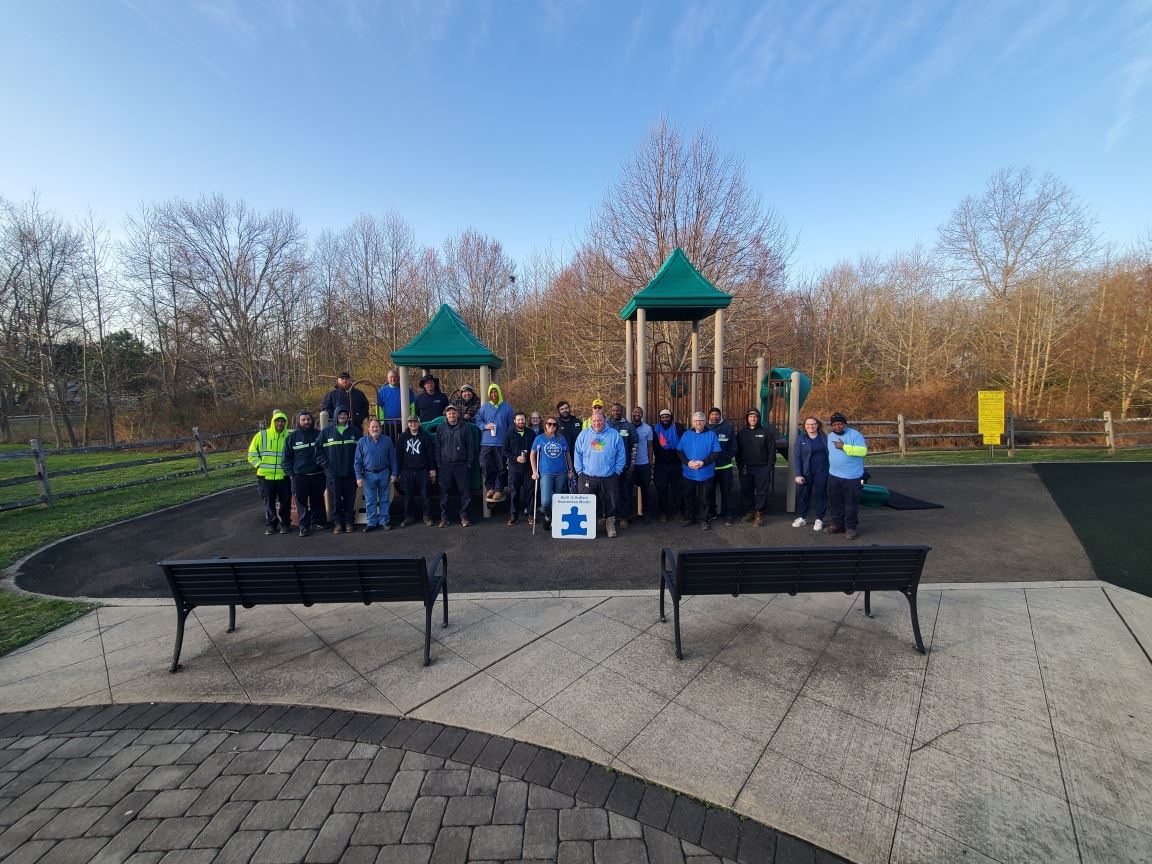 Group of people posing in a park near a green playground structure, with benches and a yellow sign at right.